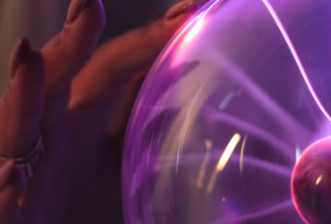 A close-up of a woman's hand touching a glass tesla coil. She is wearing rings and nail polish.