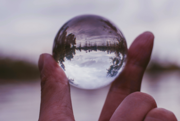 A thumb and forefinger hold a glass orb that shows sky, water and trees upside down.